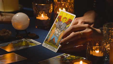 Close-Up-Of-Woman-Giving-Tarot-Card-Reading-On-Candlelit-Table-Holding-The-Lovers-Card-1